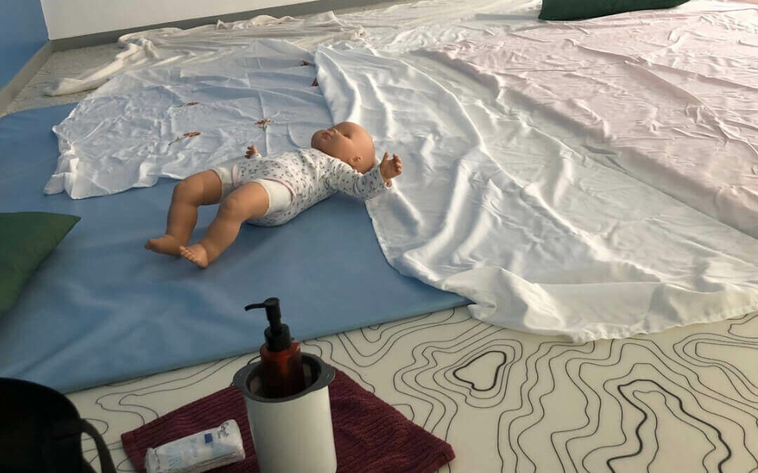 Dans une salle de la crèche près de Valenciennes, un poupon de démonstration repose sur un tapis, à côté d'un flacon d'huile végétale. L'espace est préparé pour un atelier collectif de massage bébé ayurvédique à Beuvrages