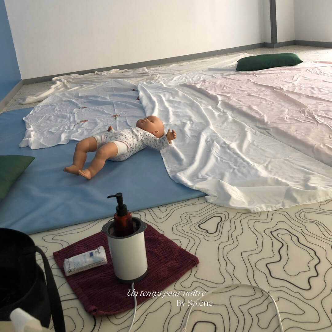 Dans une salle de la crèche près de Valenciennes, un poupon de démonstration repose sur un tapis, à côté d'un flacon d'huile végétale. L'espace est préparé pour un atelier collectif de massage bébé ayurvédique à Beuvrages
